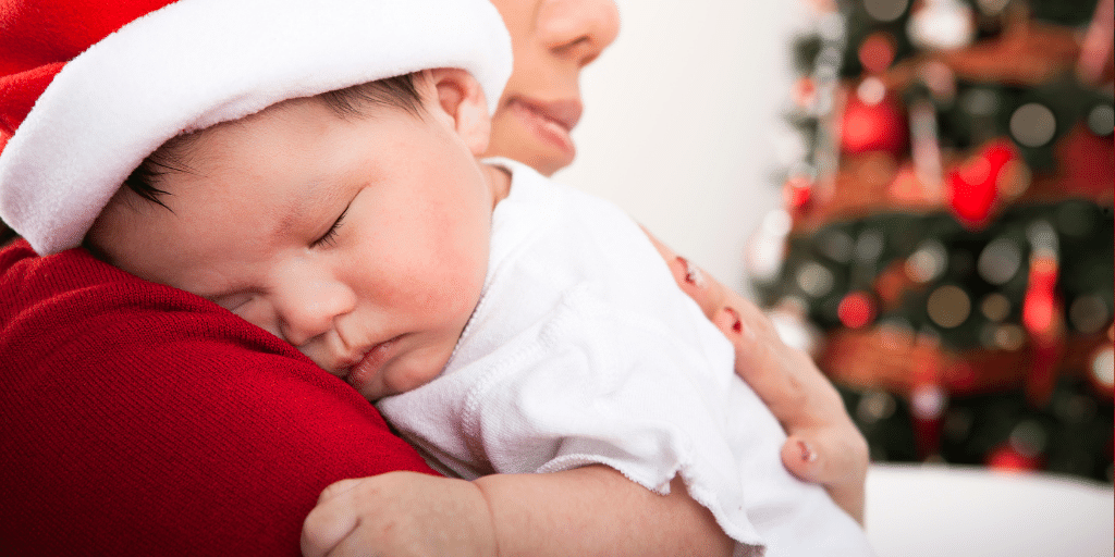 image of a baby in a christmas hat sleeping on adult with christmas tree in the background